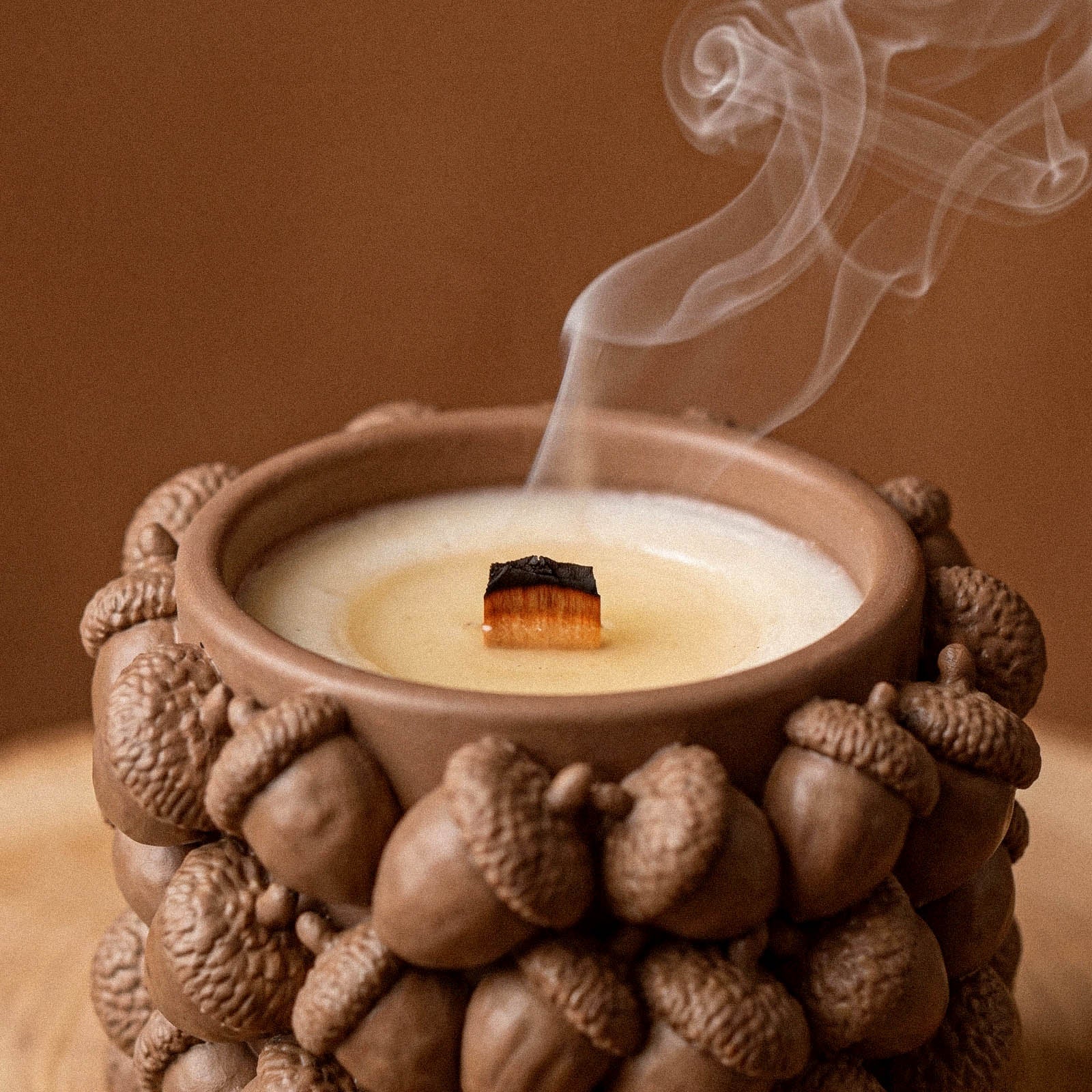 Close-up of a wooden wick candle burning inside an acorn-textured jar, with a thin curl of smoke rising.