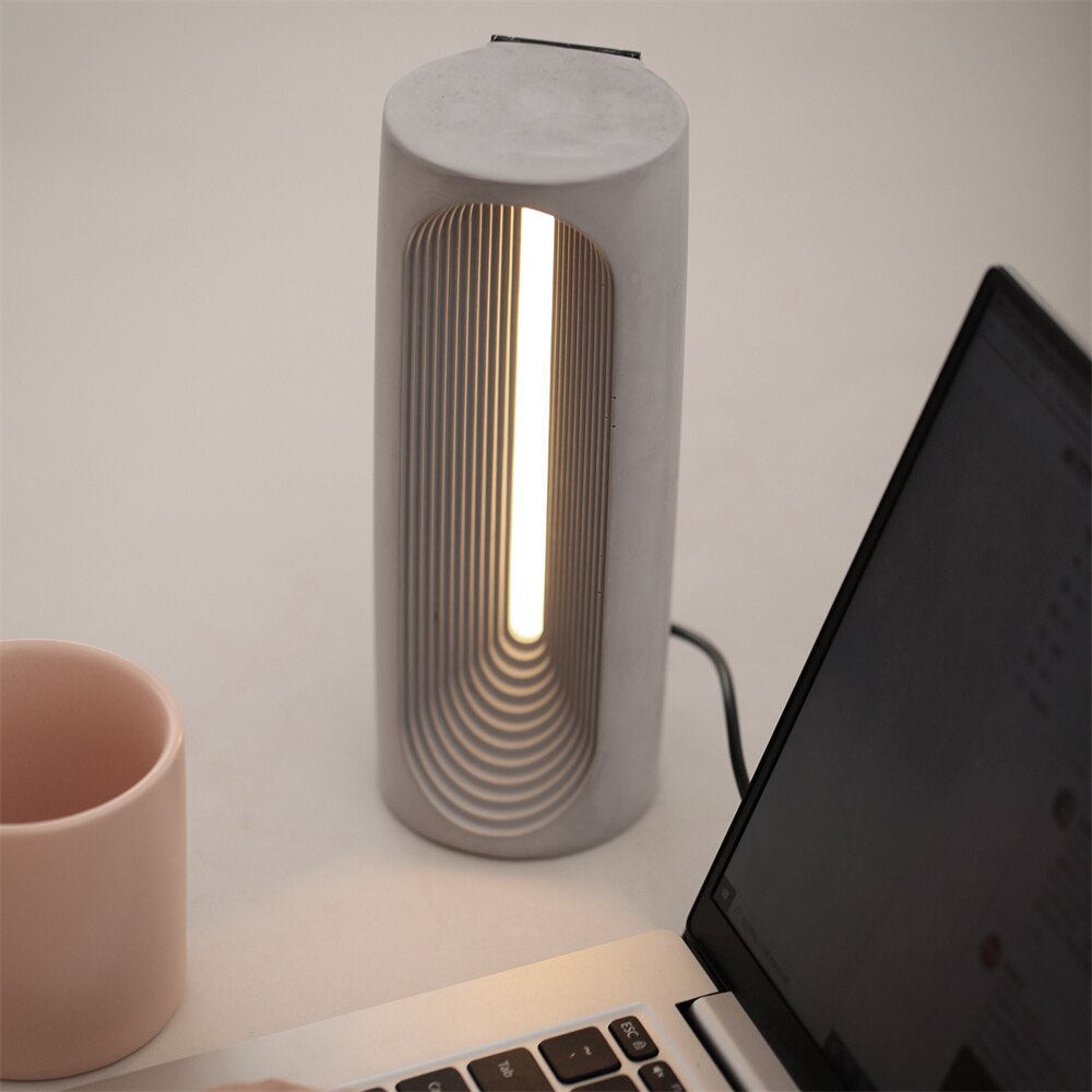 Experience the glow of productivity – a lit concrete table lamp brightens the scene next to a desktop computer, creating an ambiance of focus and warmth.